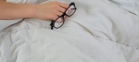 person lying on bed while covering face with pillow and holding eyeglasses